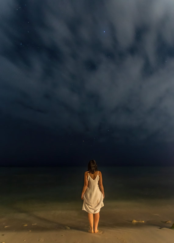 a girl in a white dress, stands at the edge of the sea watching the stars at night