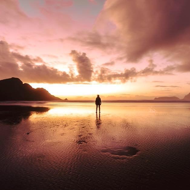 A girl stands on the endless beach looking out a t sunset over the mountains and ocean