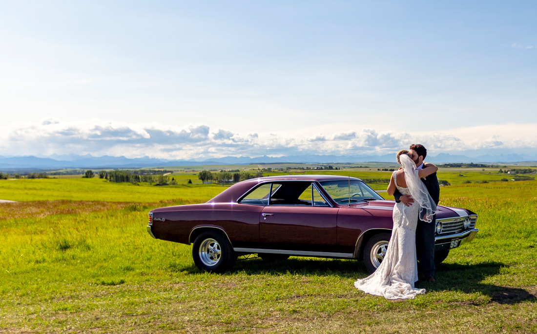 groom cuddles the bride in front a vintage car with a mountain view